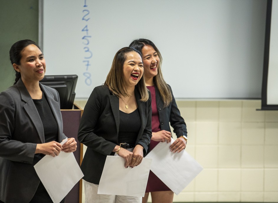 Three female students smiling and laughing while presenting an operations project in front of their class.