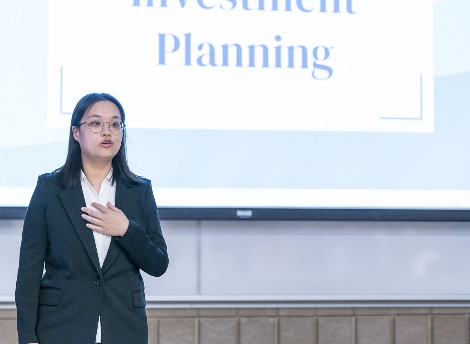 A female Asian student is standing and presenting in front of a large projection screen about Investment Planning.