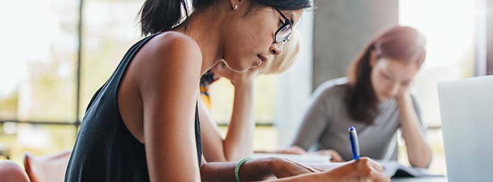 Young woman writing notes with classmates studying in background.