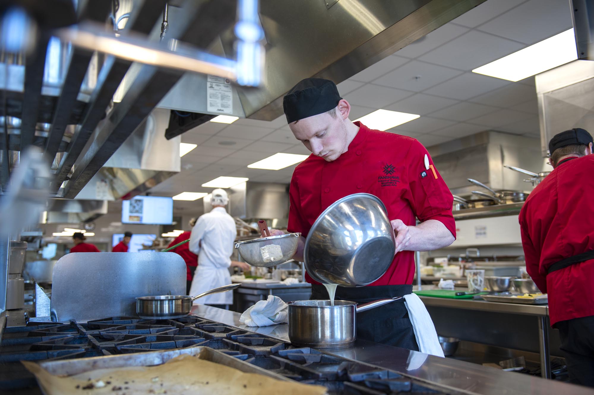 Cook working in Fanshawe College culinary labs, pouring liquid from bowl to pan on stove