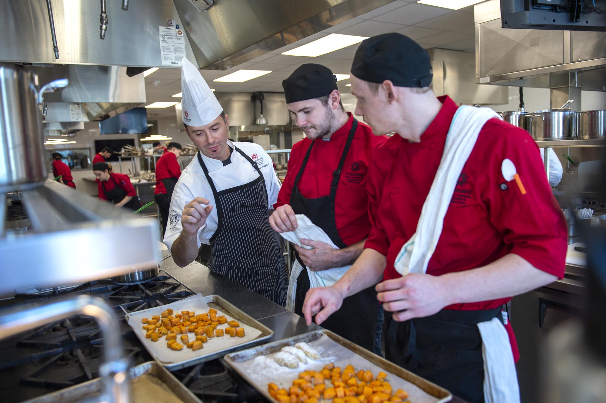 Chef instructing two students in culinary lab, standing in front of sheet pan of roasted vegetables