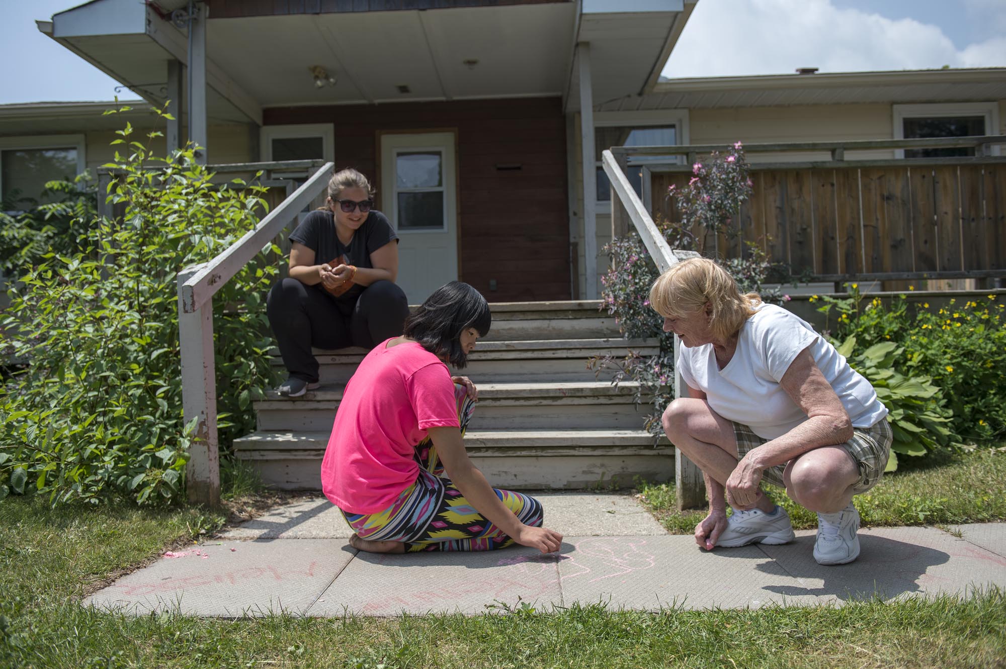 Three people sitting outside; two drawing on the concrete with chalk while another watches