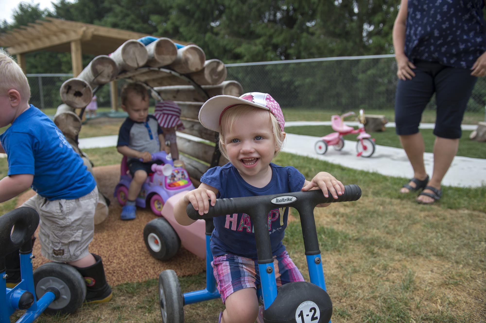 Children laughing and playing outside on tricycles while an caregiver looks on