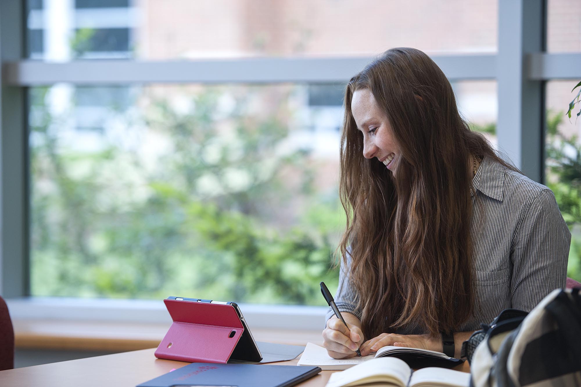 Student writing at table while looking at tablet