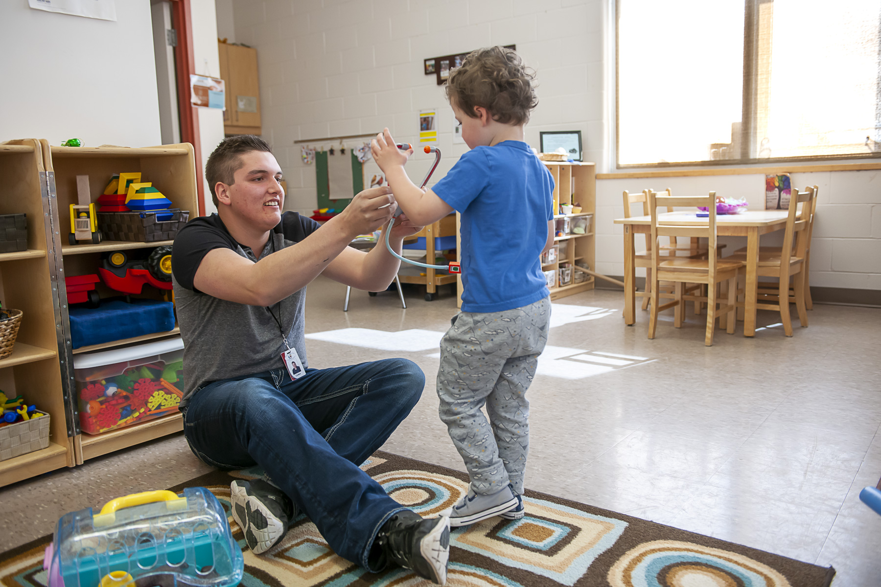 Early Childhood educator sits on the floor interacting with a toddler in a classroom