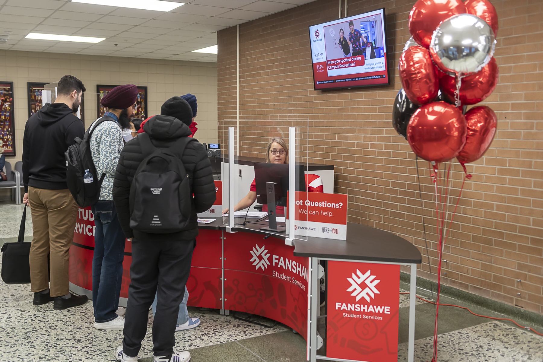men stand at a desk talking to a woman behind the desk next to some balloons