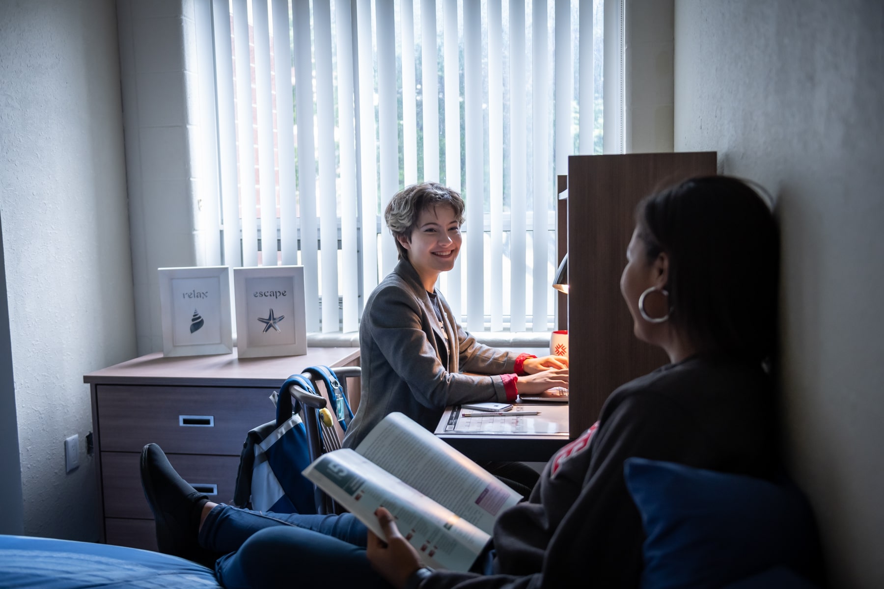 Woman doing homework smiling at roommate who is sitting on bed