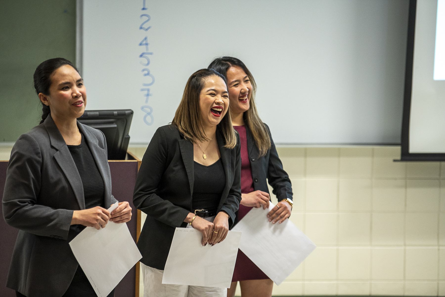 Three female students smiling and laughing while presenting an operations project in front of their class.