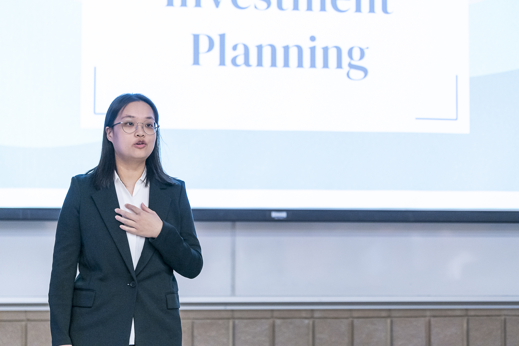 A female Asian student is standing and presenting in front of a large projection screen about Investment Planning.