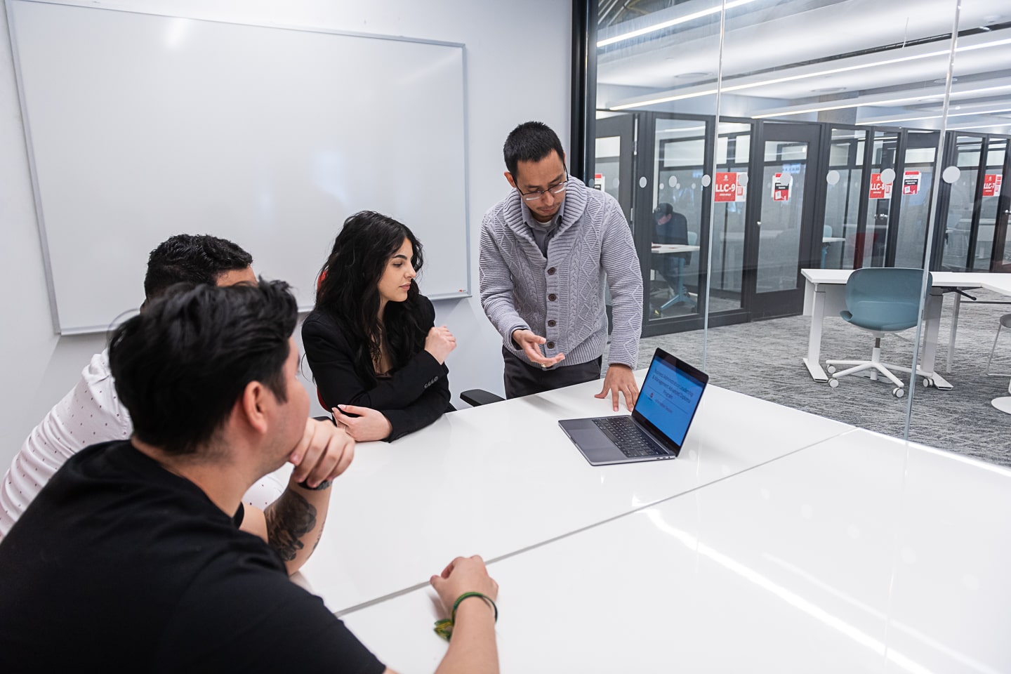 man standing points to laptop on table while three people watch