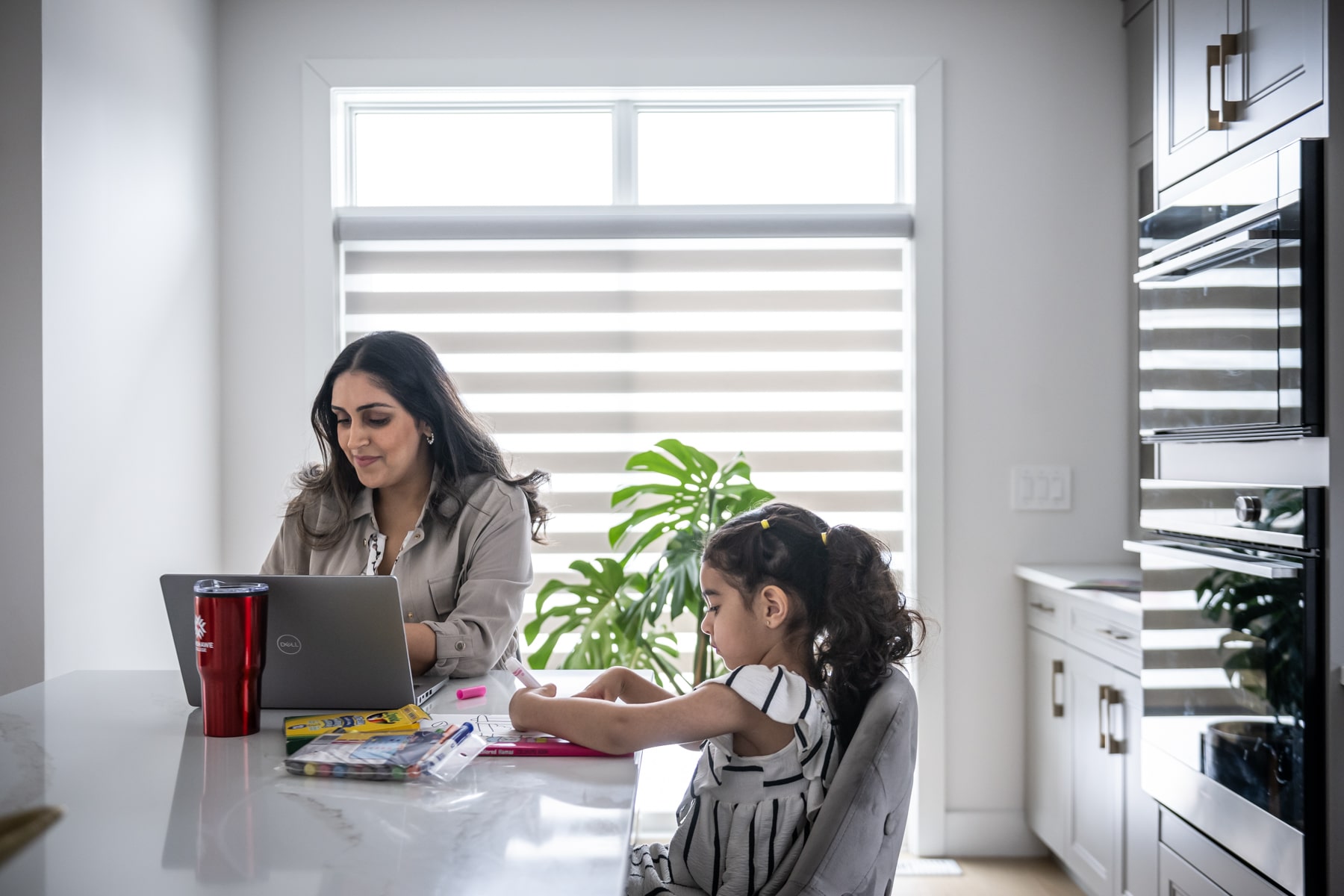 mother sits at laptop studying while daughter draws beside her