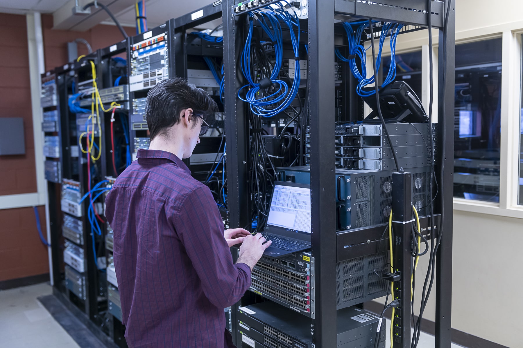man stands in front of servers while typing on a laptop
