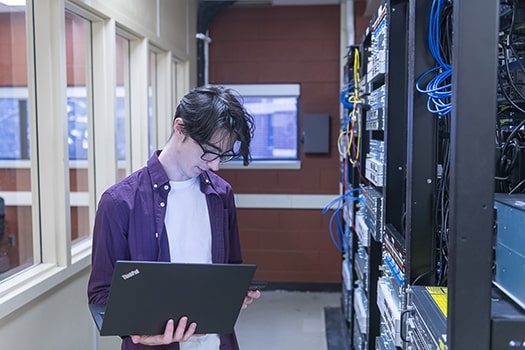 Information Systems Specialist man stands beside server holding a laptop