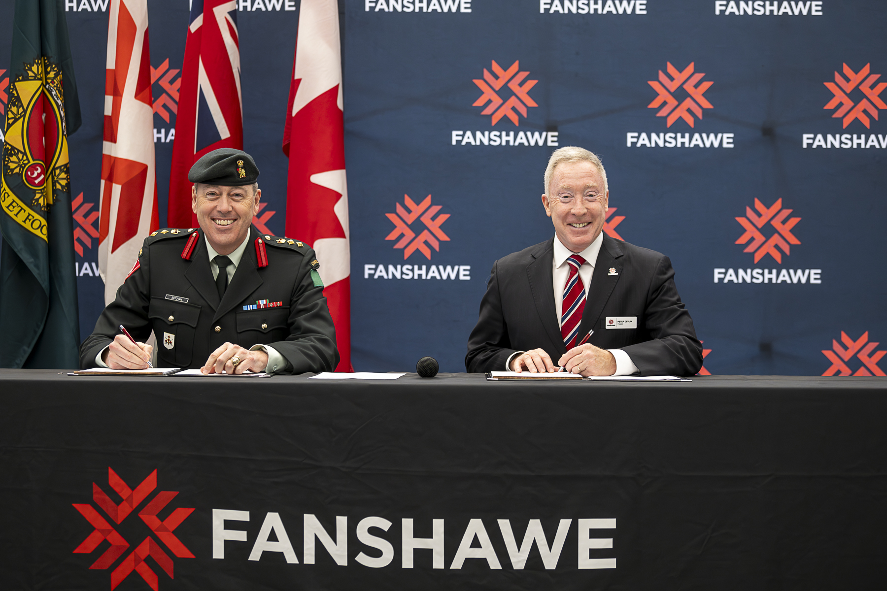 Colonel Chris Brown and President Peter Devlin seated at a table in front of Fanshawe backdrop to sign an MOU agreement.