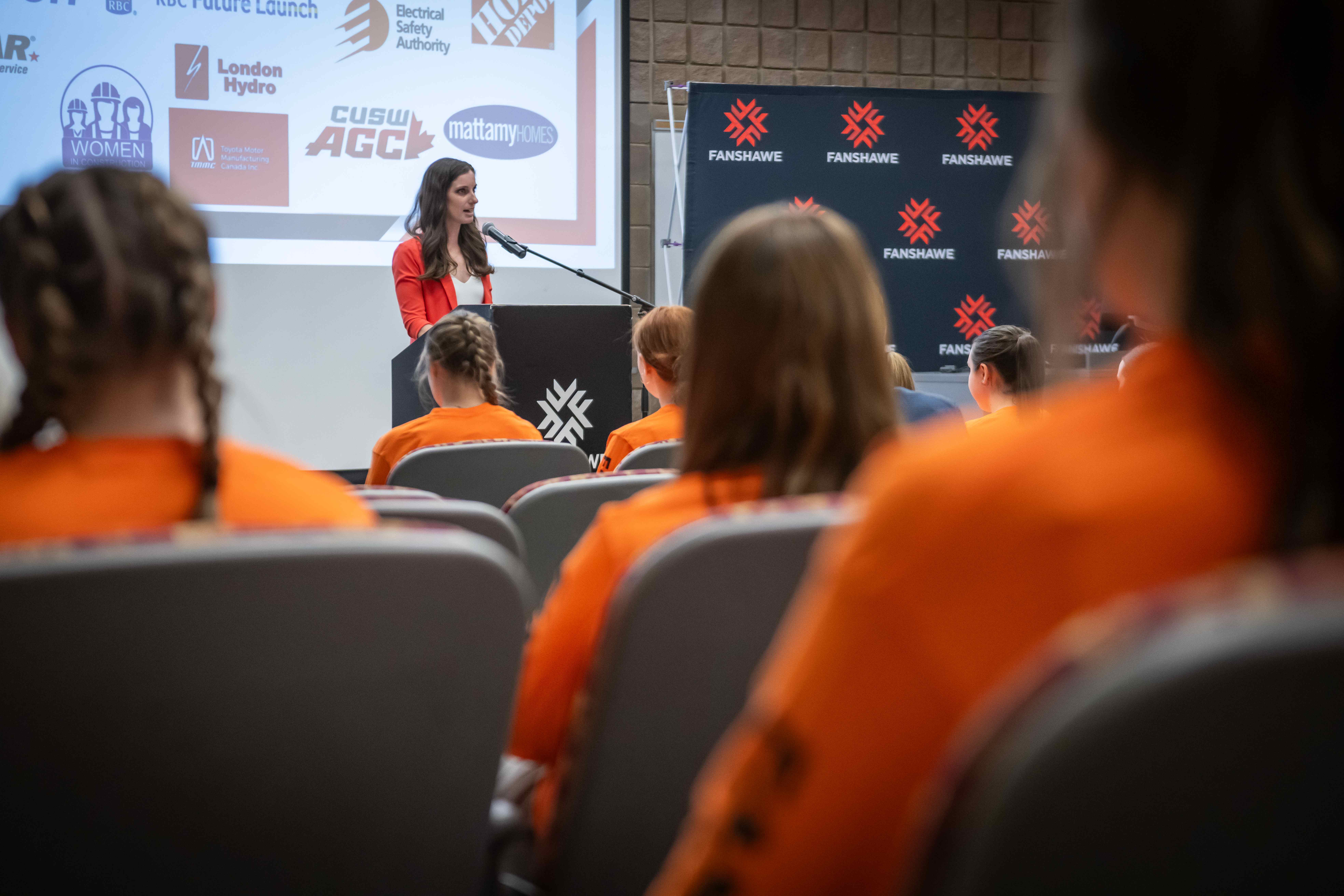 A woman stands at a podium speaking to female high school students wearing orange shirts.