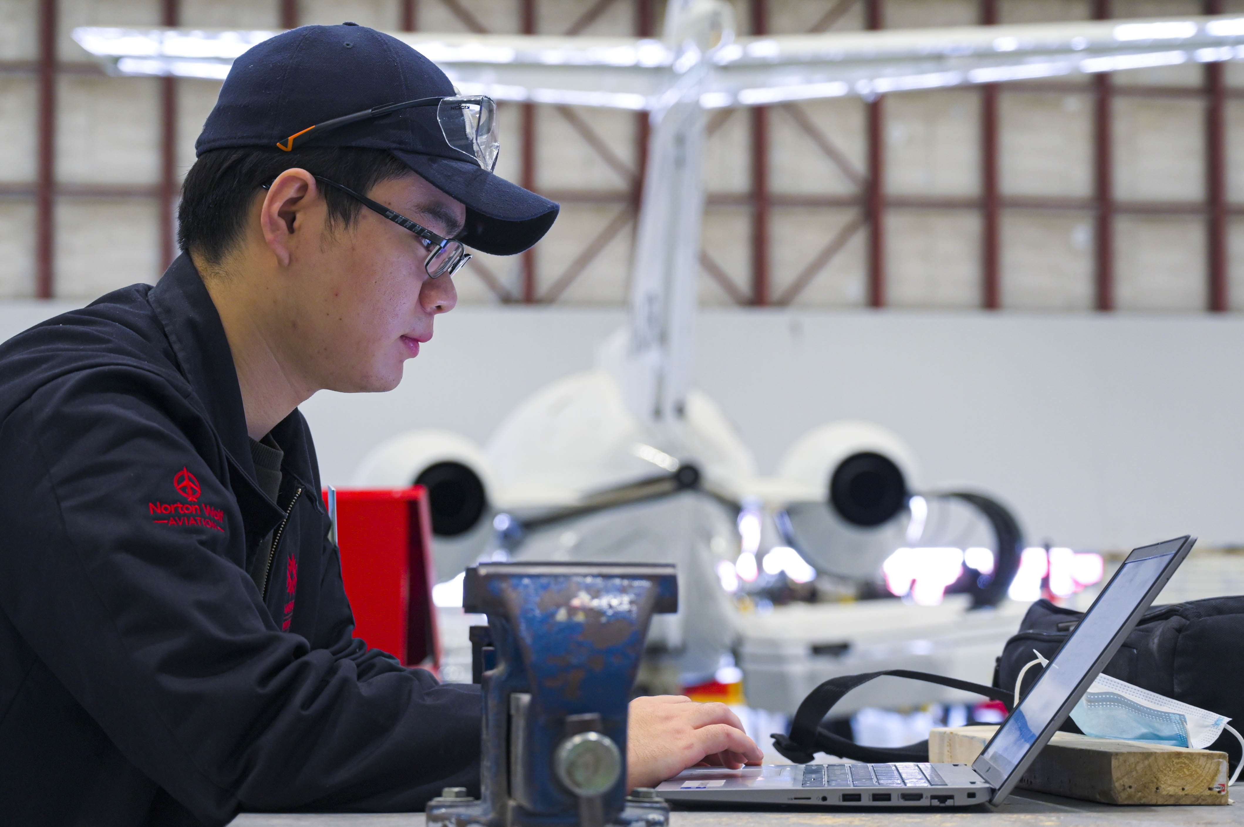 Aviation student sitting at a laptop with a plane in the background.