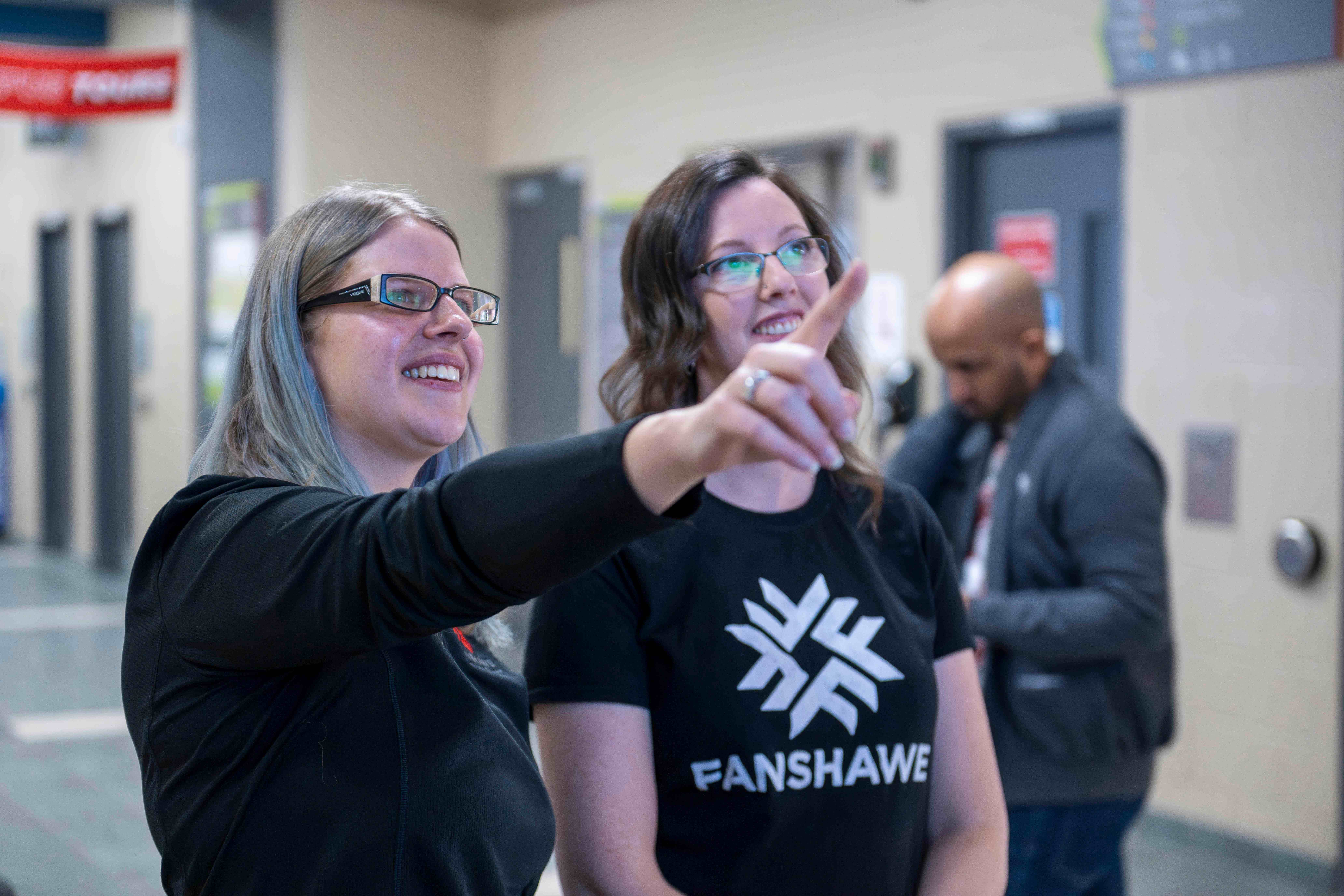 two people stand together at Fanshawe, pointing to a tv above them