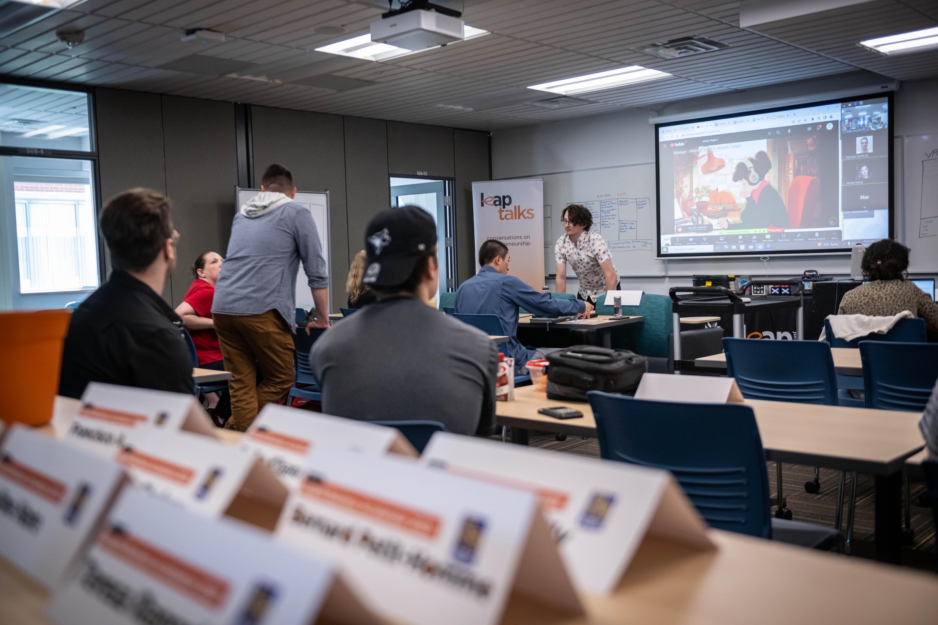 A classroom setting with tables and blue chairs, a projector screen displaying a video call, and a whiteboard with notes.
