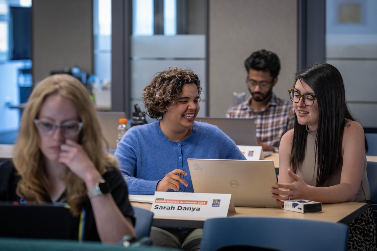 Several people sit at tables in a classroom setting, working on laptops and engaging in conversation.