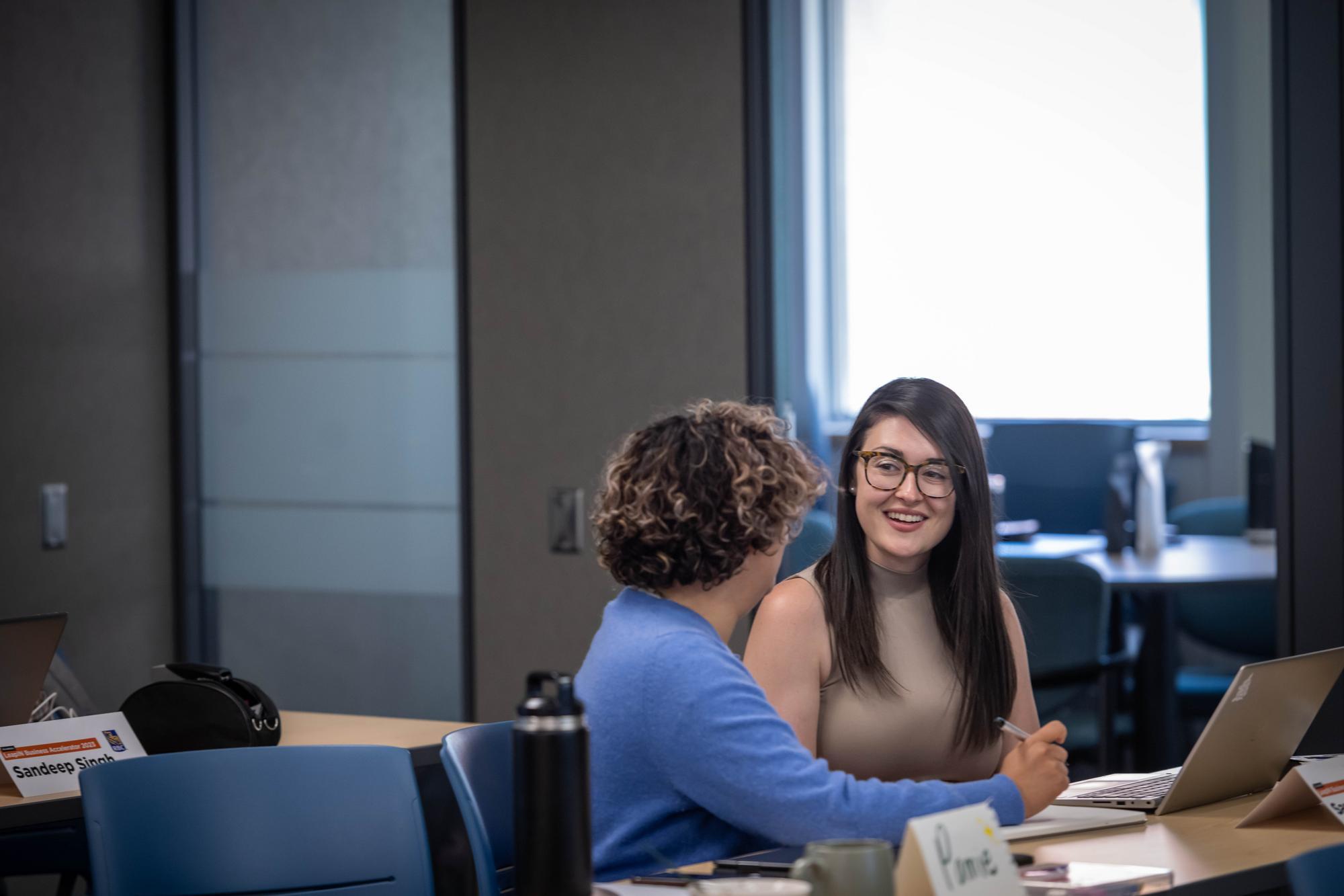 Two individuals sit at a table with laptops and notebooks, engaged in conversation.