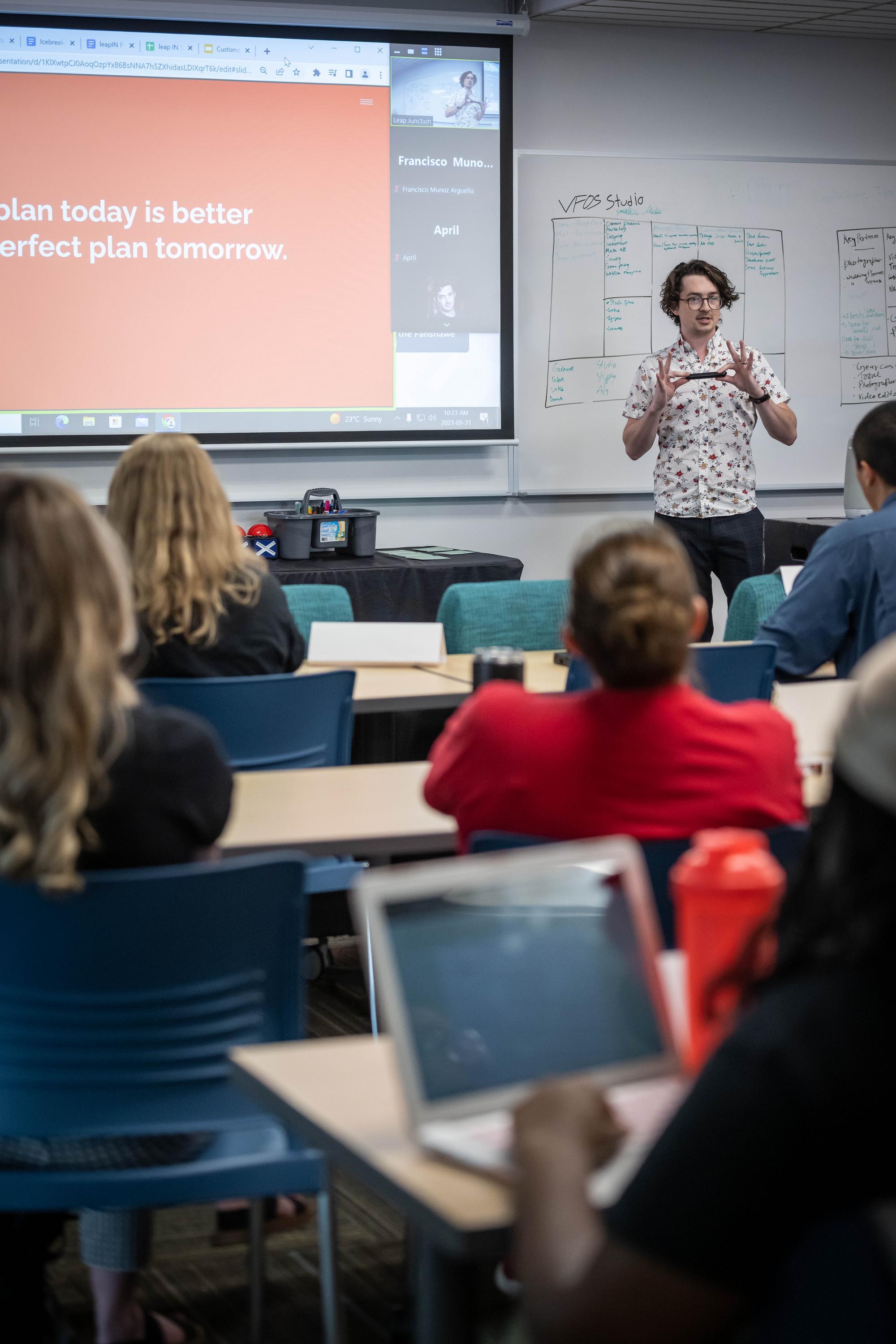 In a classroom setting, an individual stands near a whiteboard covered in notes, gesturing towards a projected screen displaying text, while others sit at desks with laptops.