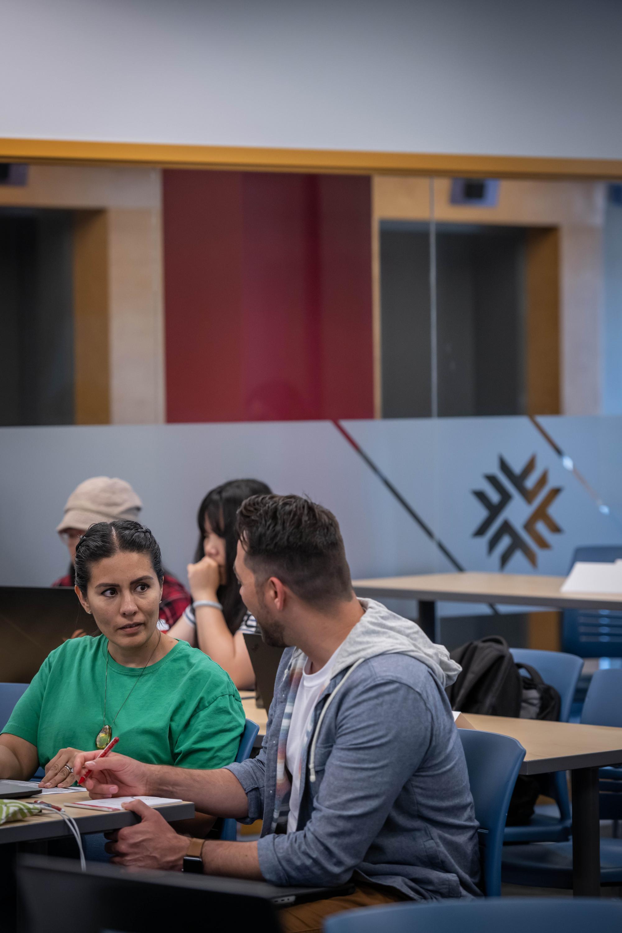 Two people sit at a table, one in a green shirt and the other in a grey hoodie, engaged in conversation.