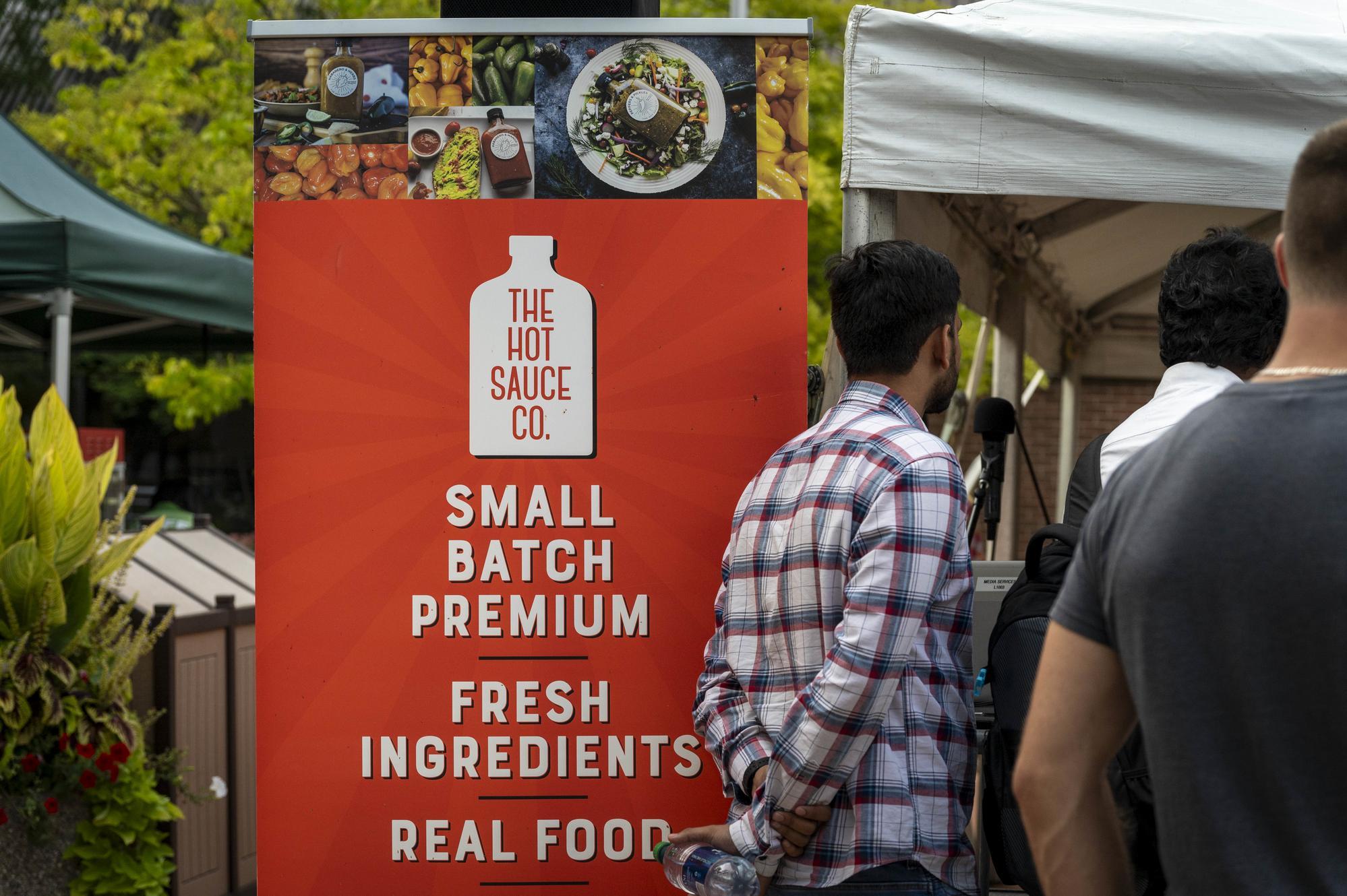 A bright orange sign advertises hot sauce with white text and a white bottle outline, with images of food and peppers above the sign.