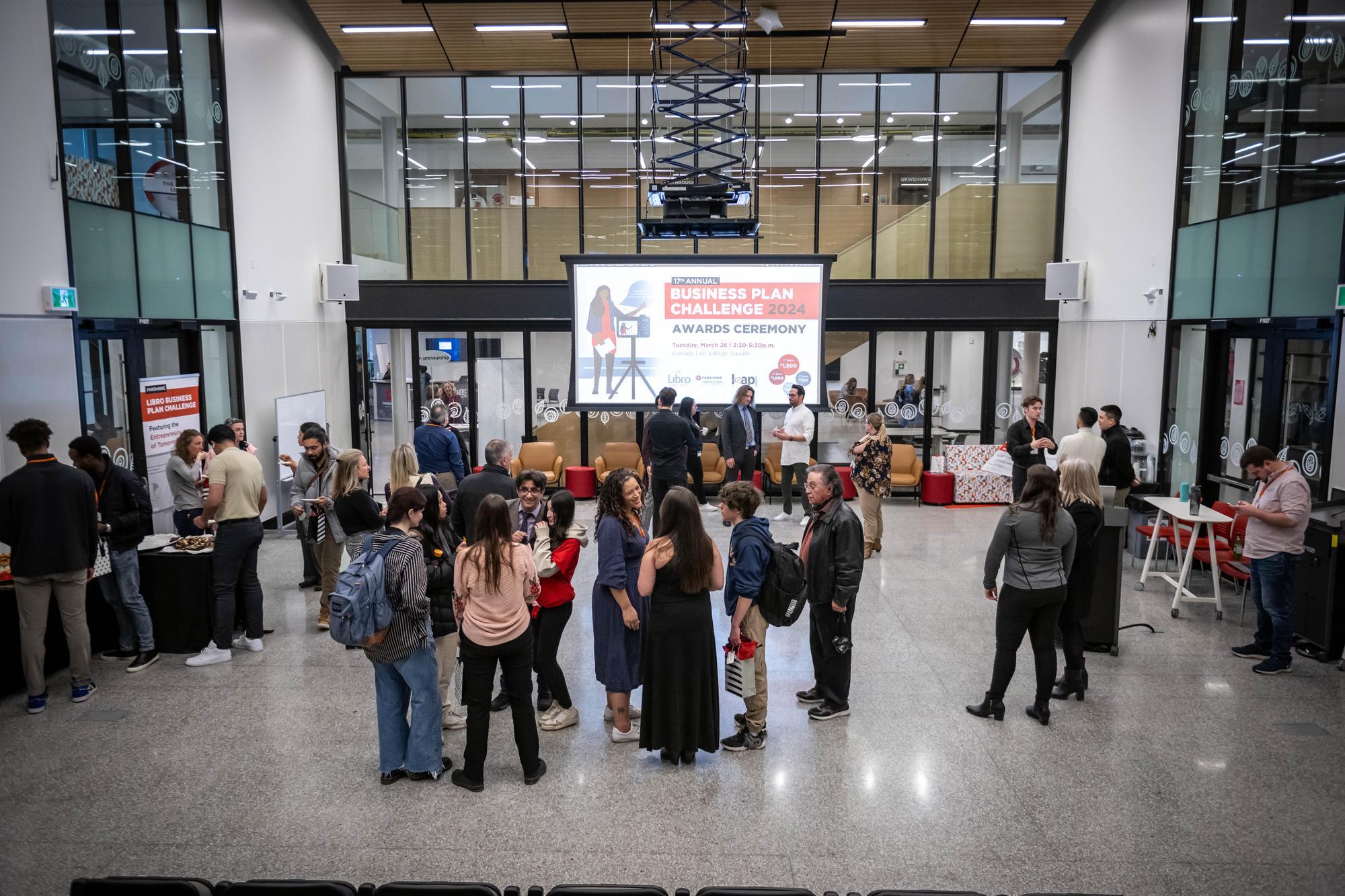 A large group of people are gathered in a spacious hall with a high ceiling and large windows, with a screen displaying information about a business plan challenge awards ceremony.