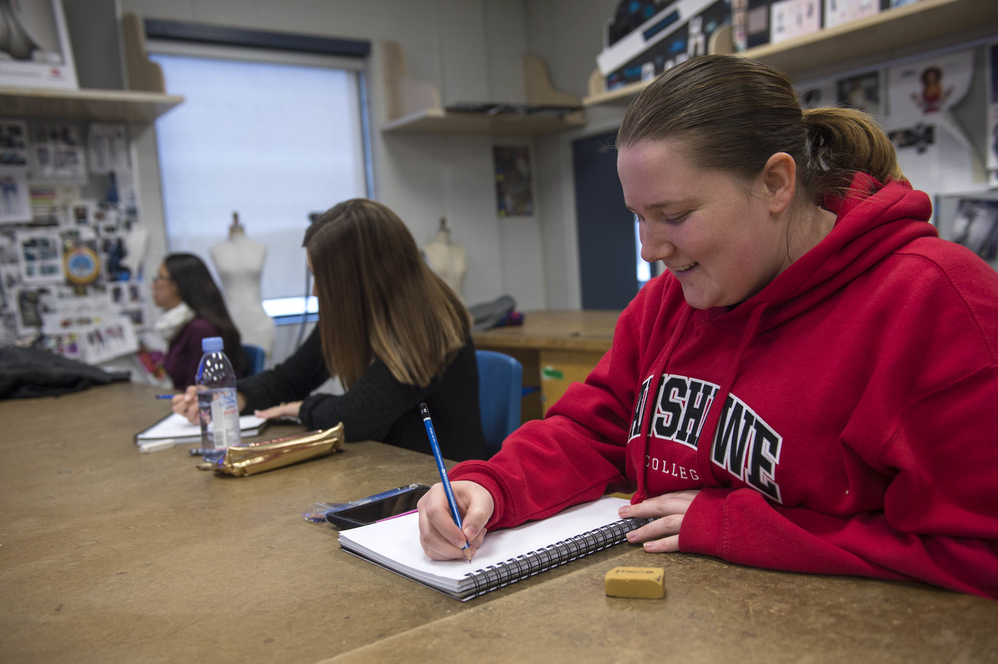 At the London Campus of Fanshawe College, three people sit at a long table, writing in notebooks with pencils, in a Design Foundations program classroom.