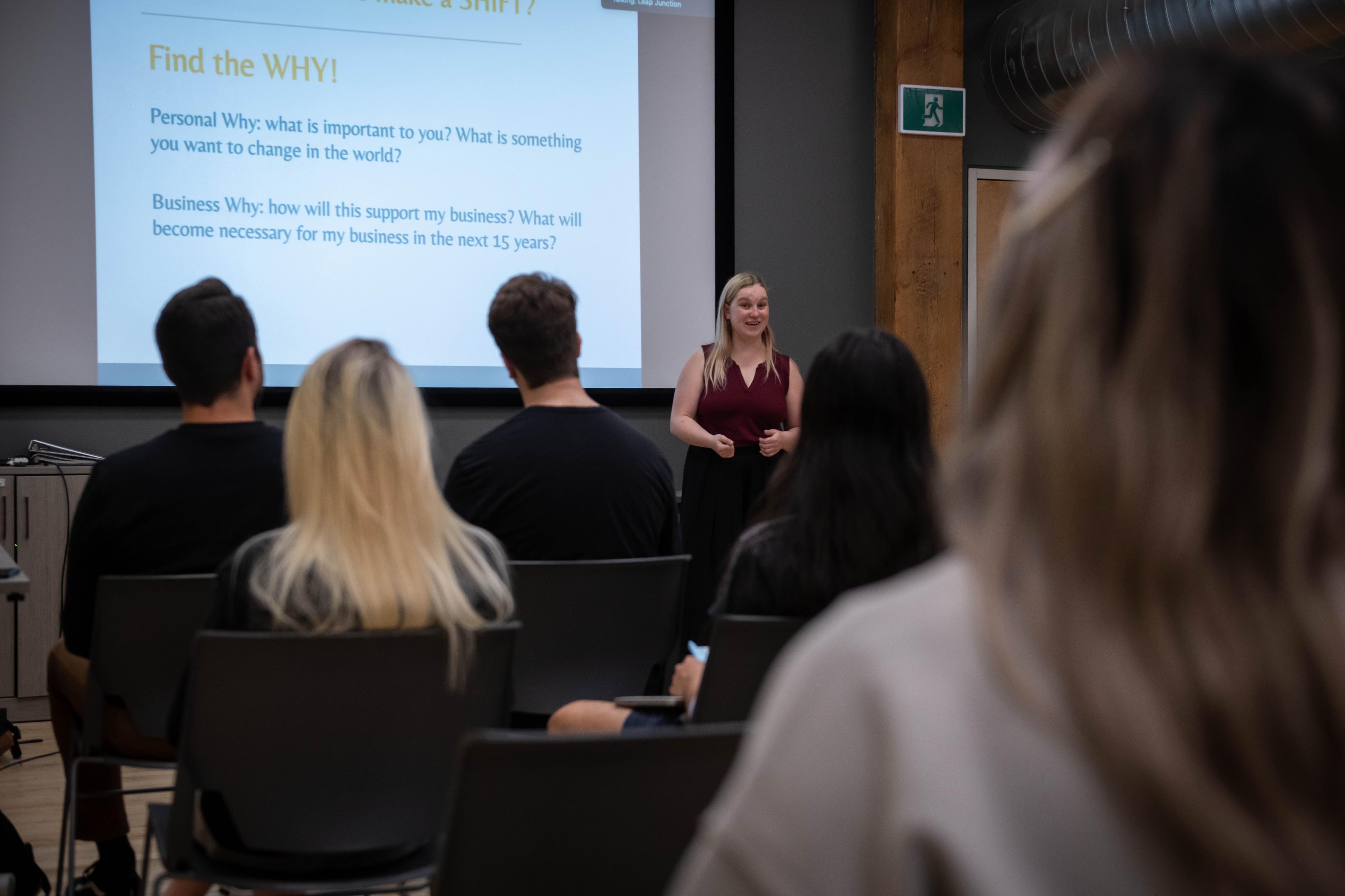 A presenter stands in front of a screen displaying text, facing an audience seated in chairs.