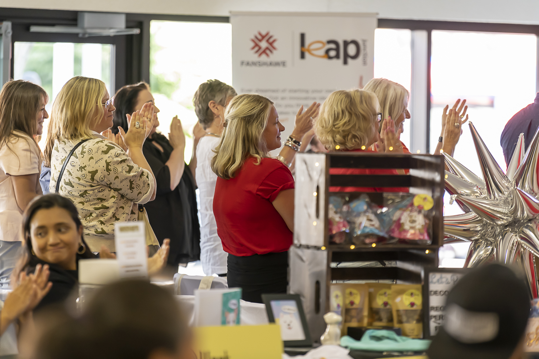 A group of people are clapping, with a banner and a display of items visible in the foreground.