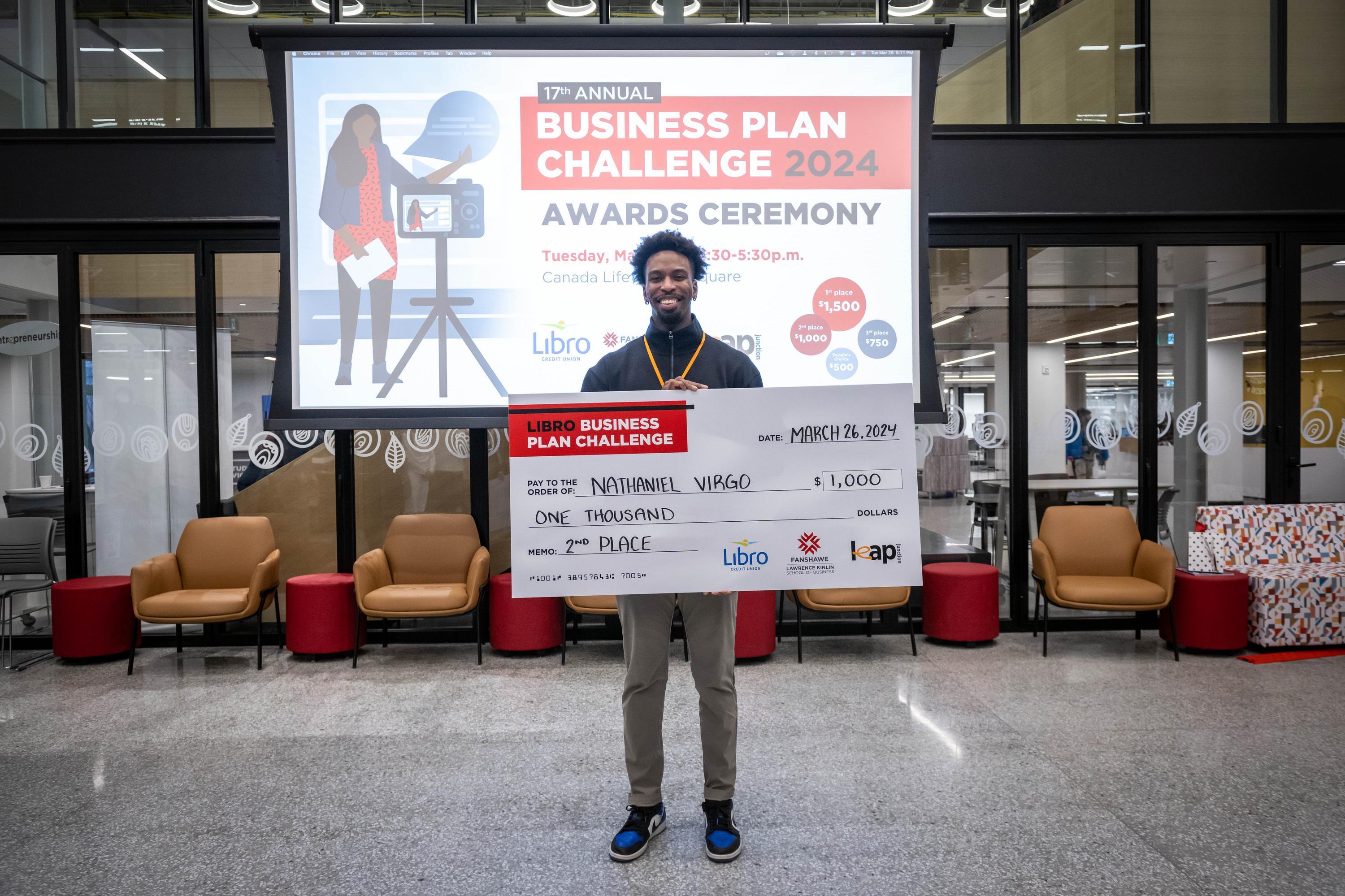 A person stands behind a podium with a laptop and microphone, presenting in front of a projected image of a greenhouse interior.
