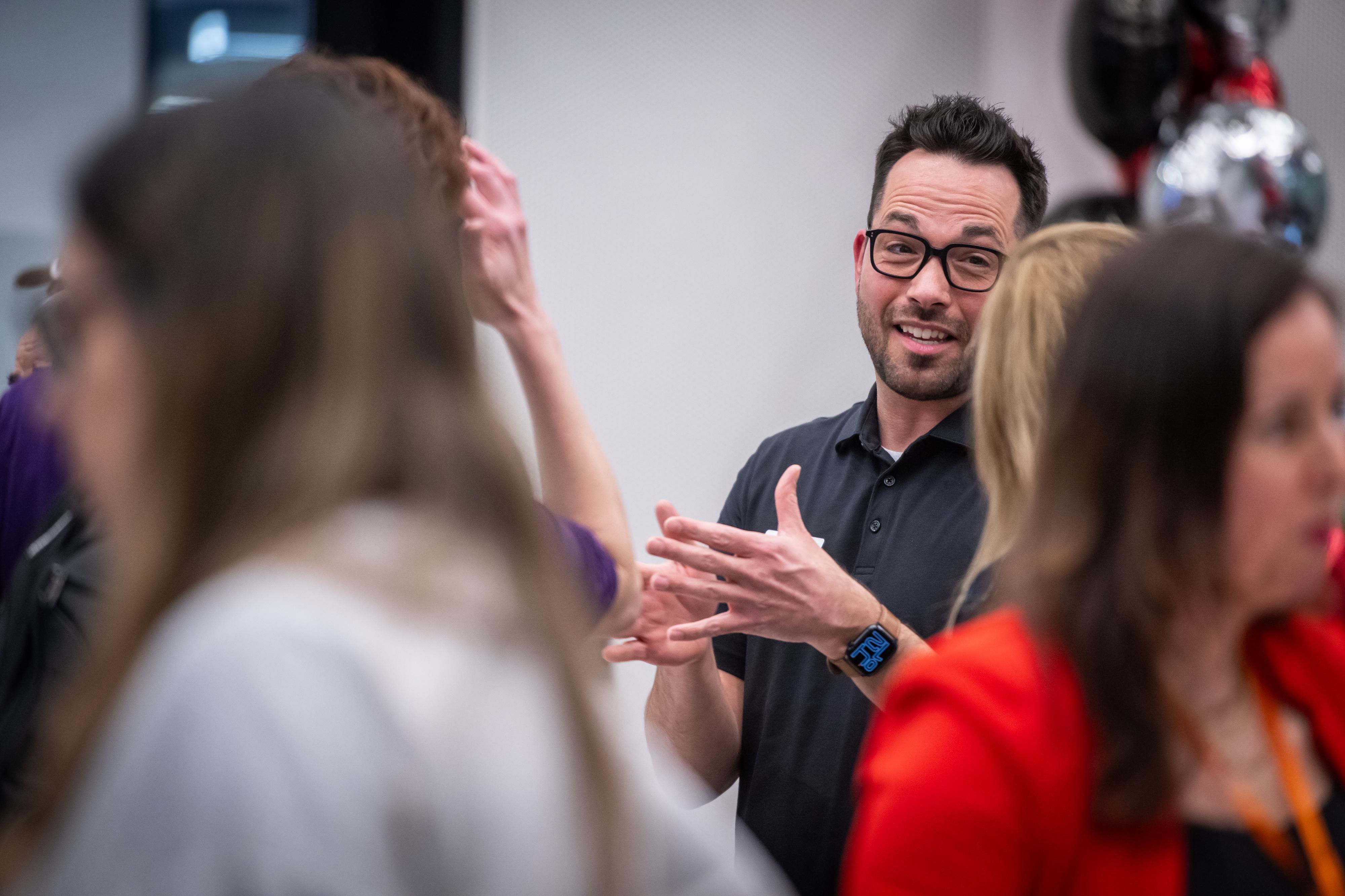 A person with glasses gestures with open hands while surrounded by others.