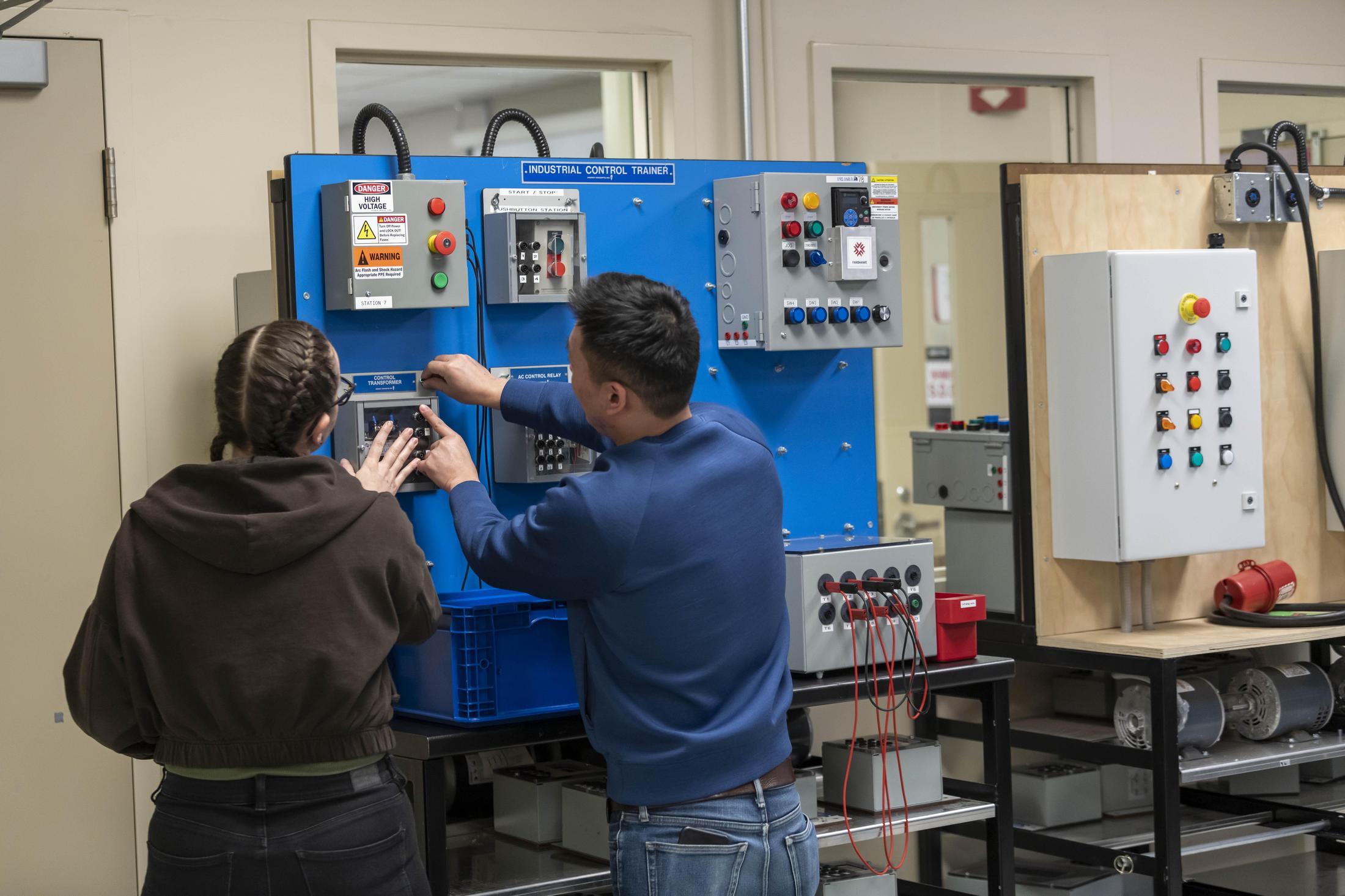 Two people stand in front of an Industrial Control Trainer, which is a blue board with various electrical components and warning signs attached.