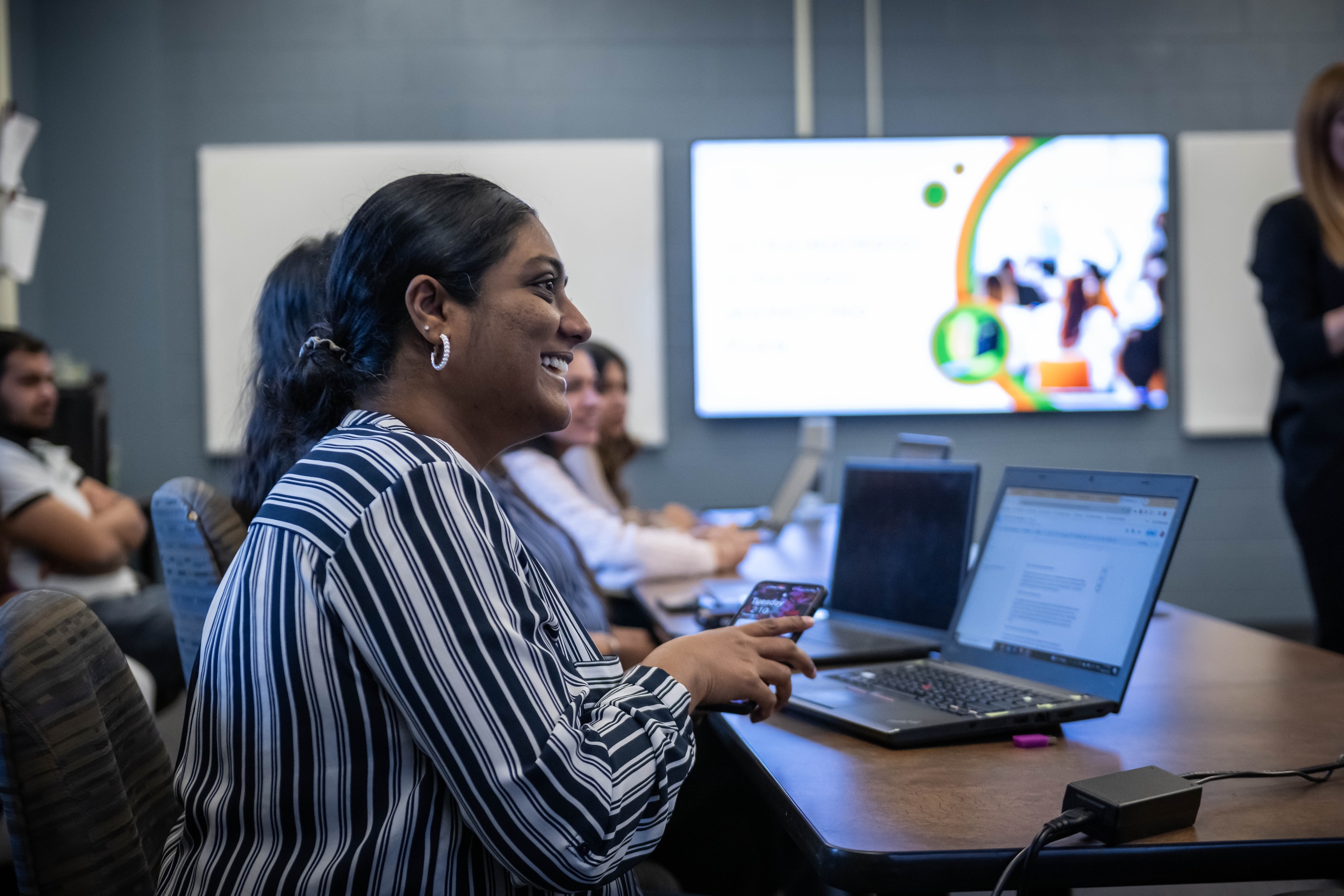 At Fanshawe College's London Campus, a business class is in session, with a person in a striped shirt smiling while holding a phone, surrounded by laptops on a table, and a presentation