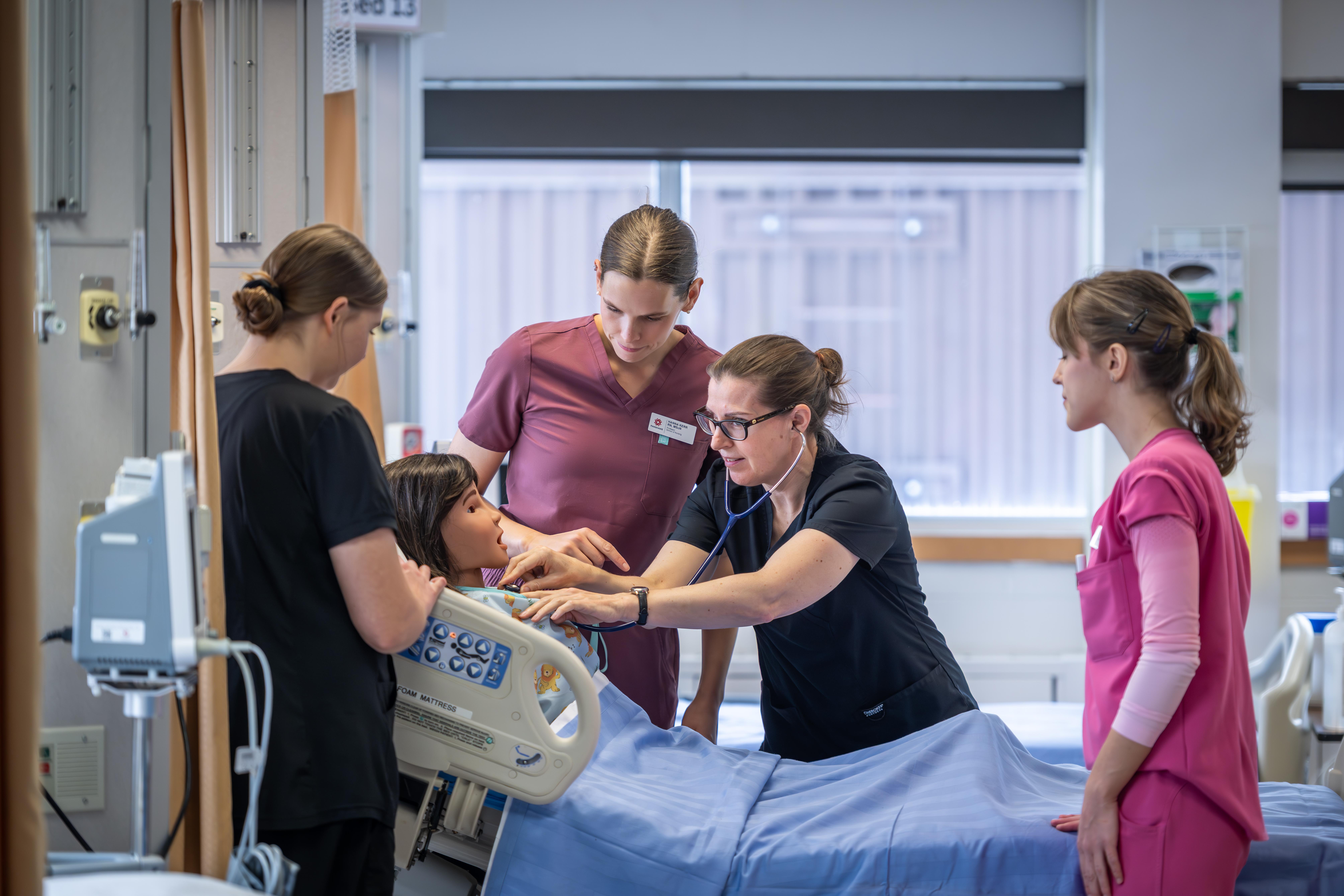 Four individuals in medical scrubs surround a patient simulator in a hospital room, one using a stethoscope.