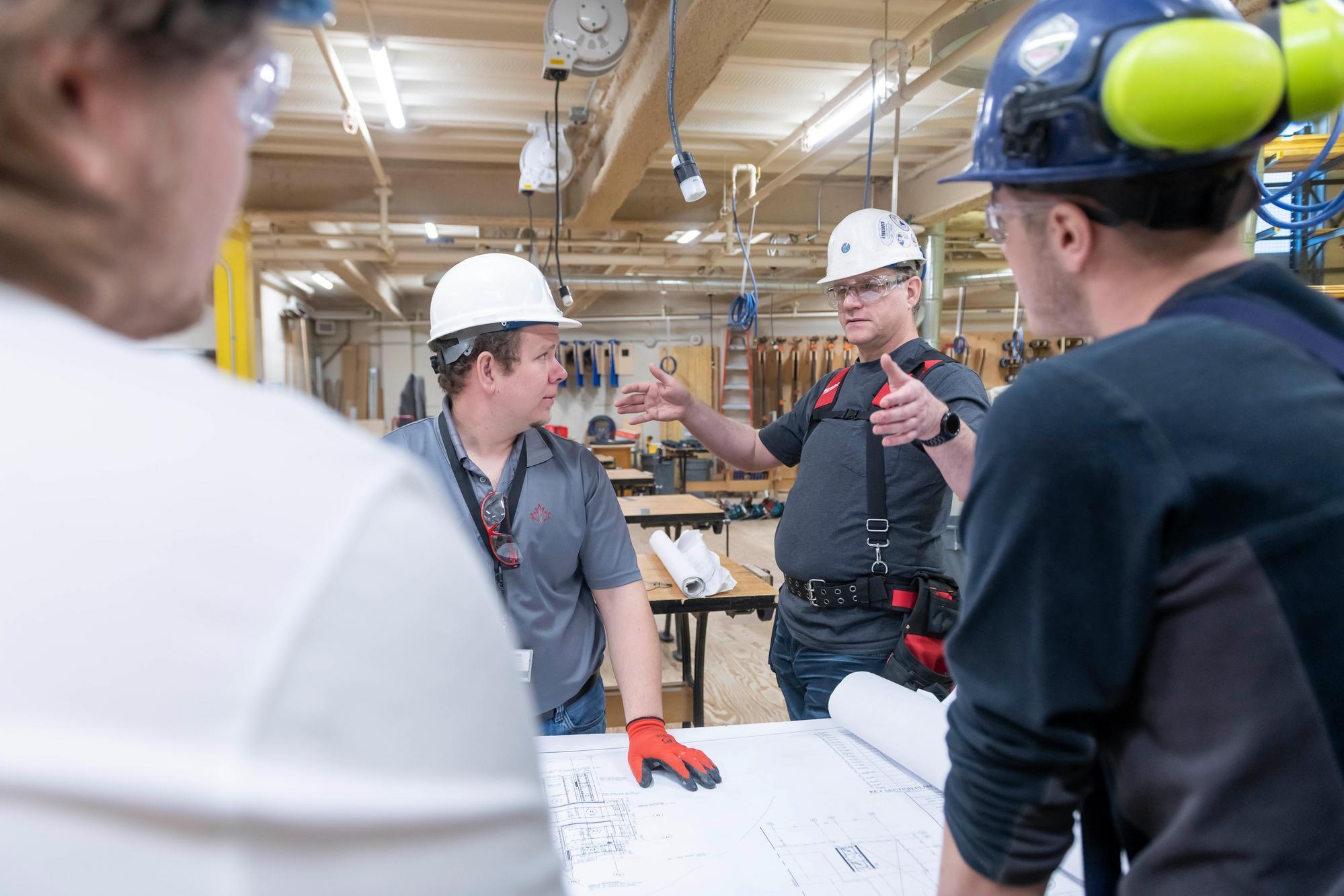 In a Fanshawe College workshop, individuals wearing hard hats and safety glasses examine a large blueprint spread across a table.