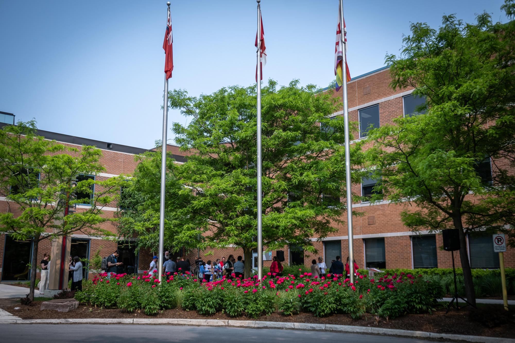 A red-brick building with large windows is surrounded by green trees and vibrant red flowers. Three flagpoles stand prominently, with a group of people gathered nearby.
