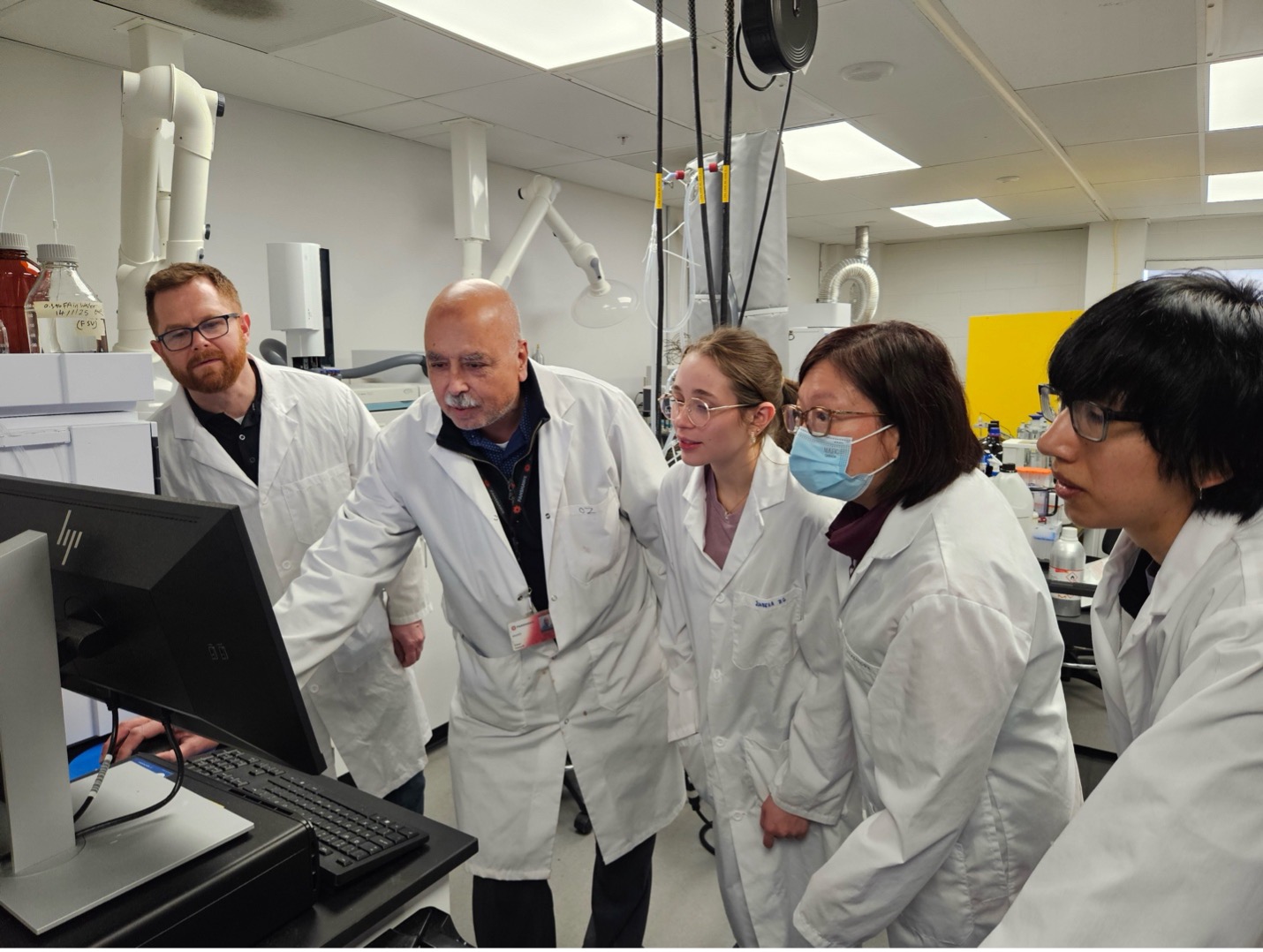 A group of five lab technicians in white coats gather around a computer in a bright laboratory. They appear focused and engaged, analyzing data on the screen.
