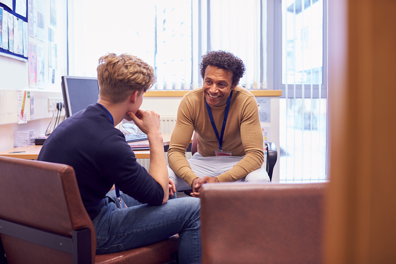 A counselor and a young man sit in an office talking. The counselor smiles warmly, creating a supportive and empathetic atmosphere.