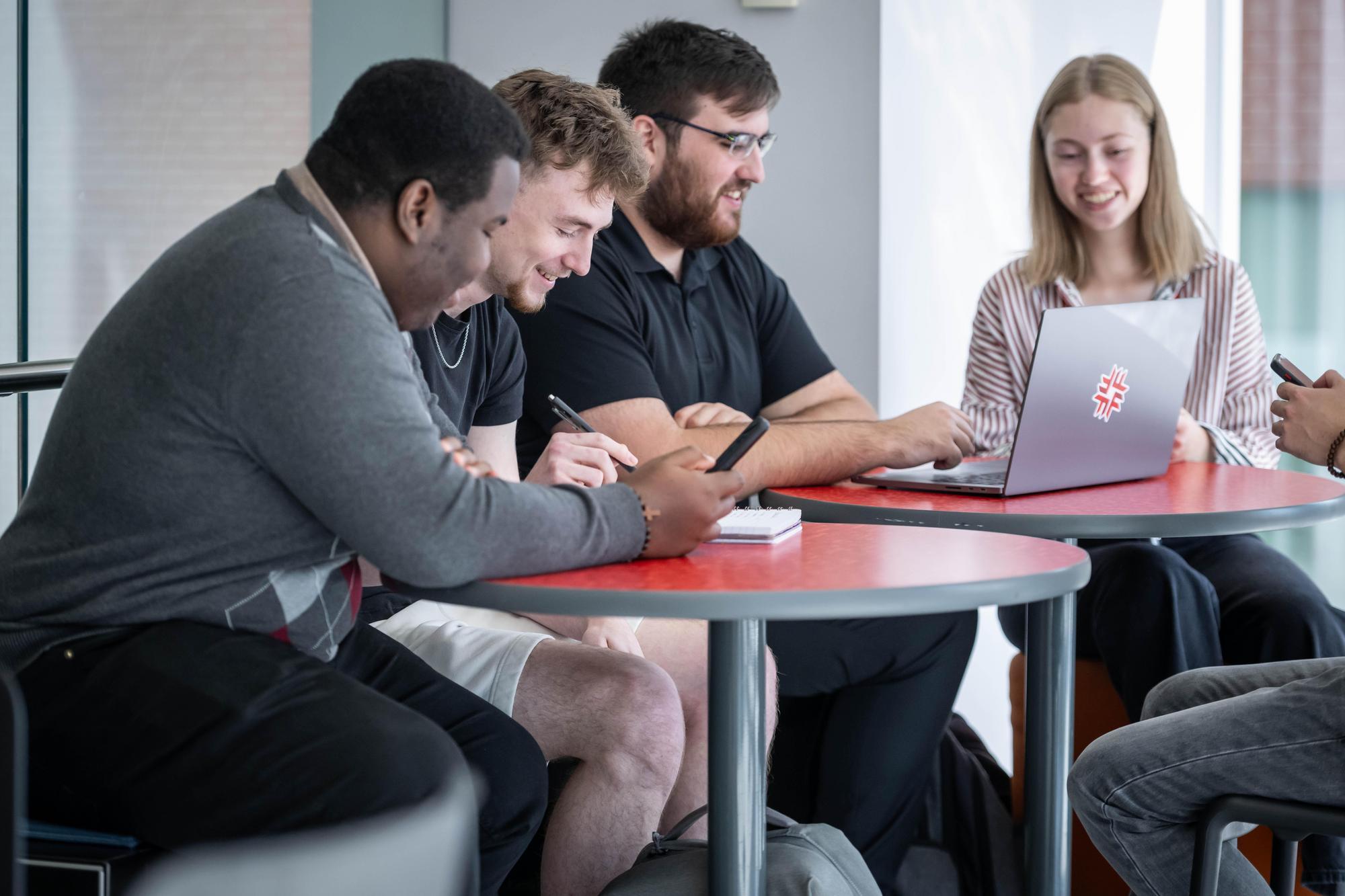 A group collaborates at a red table with a laptop bearing the Fanshawe College logo, inside the London Downtown Campus.