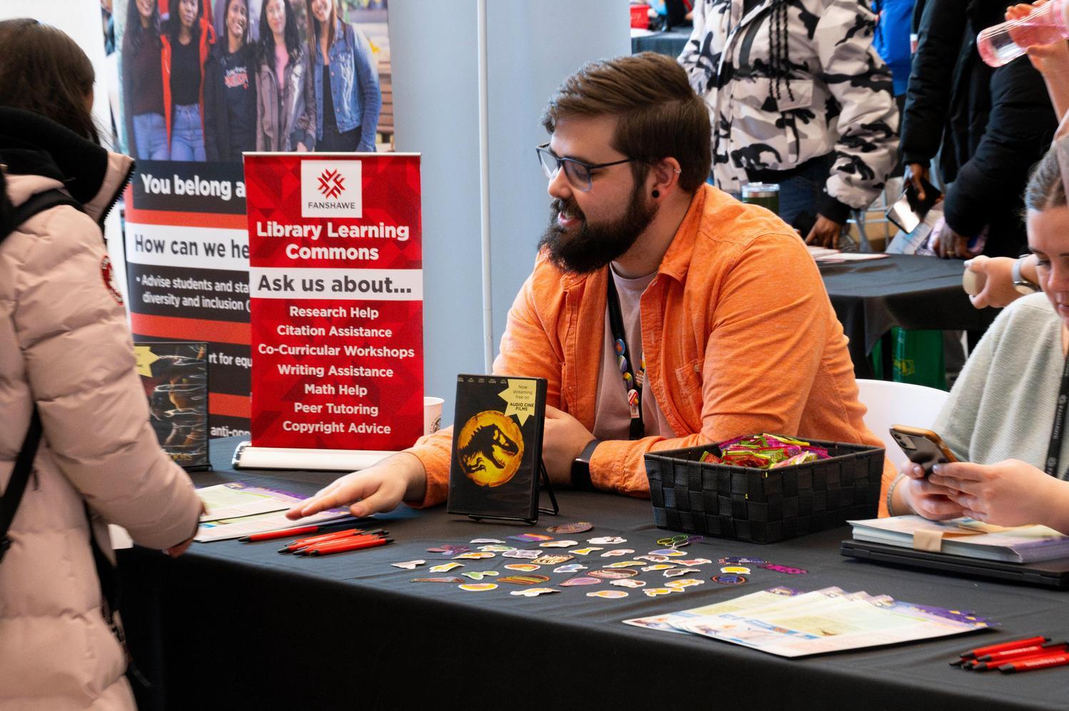 A man in an orange shirt sits at a table covered with colorful pins and brochures. A red sign reads "Library Learning Commons." The atmosphere is interactive.