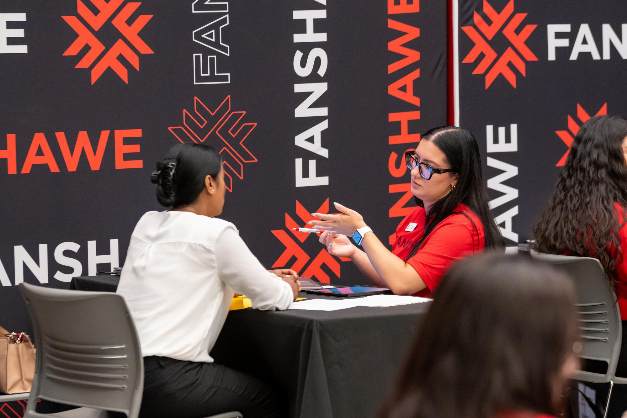 Two individuals sit at a table with a black tablecloth, in front of a backdrop with a repeating pattern