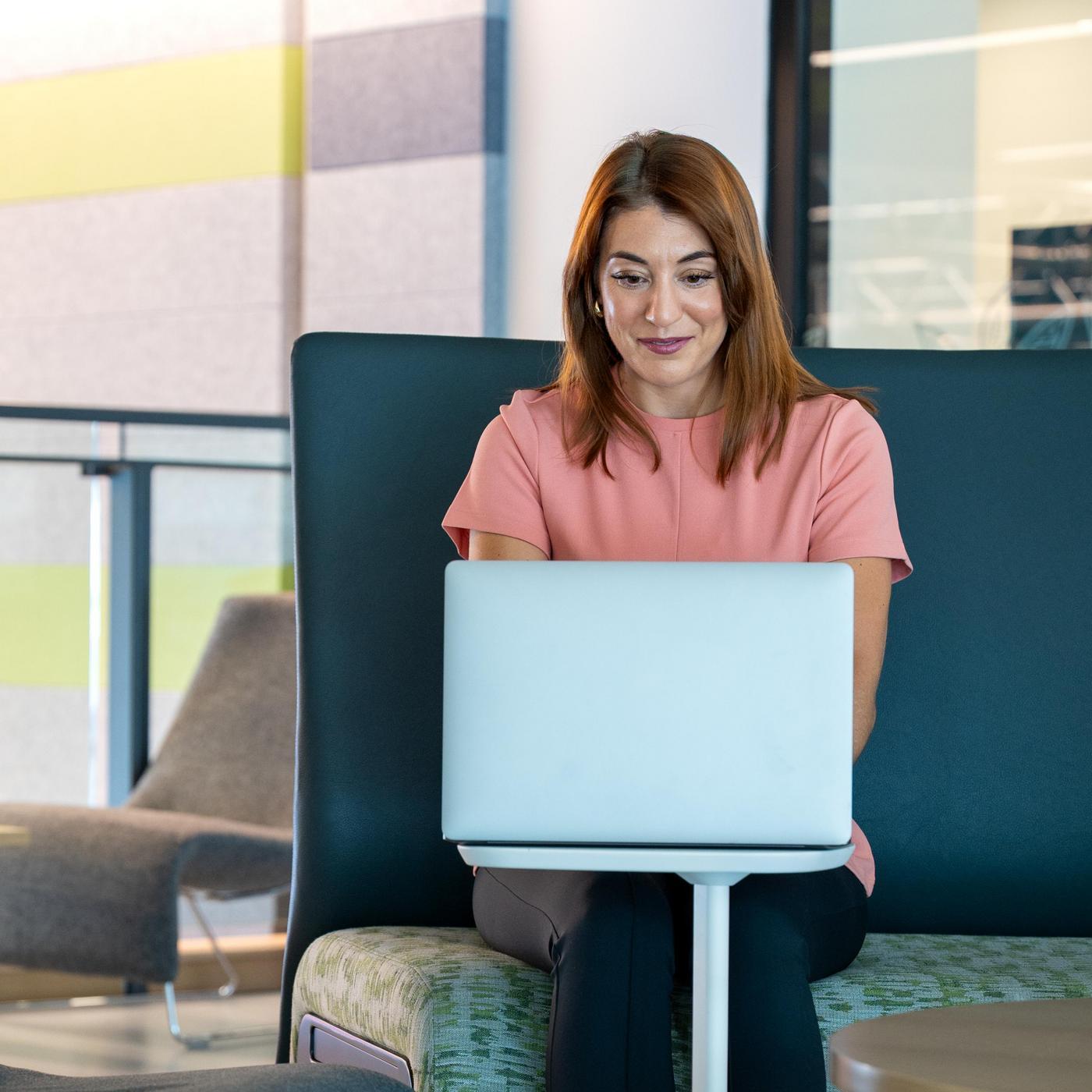 A woman with brown hair works on a laptop, sitting on a modern couch with a focused expression. The setting is bright and contemporary.