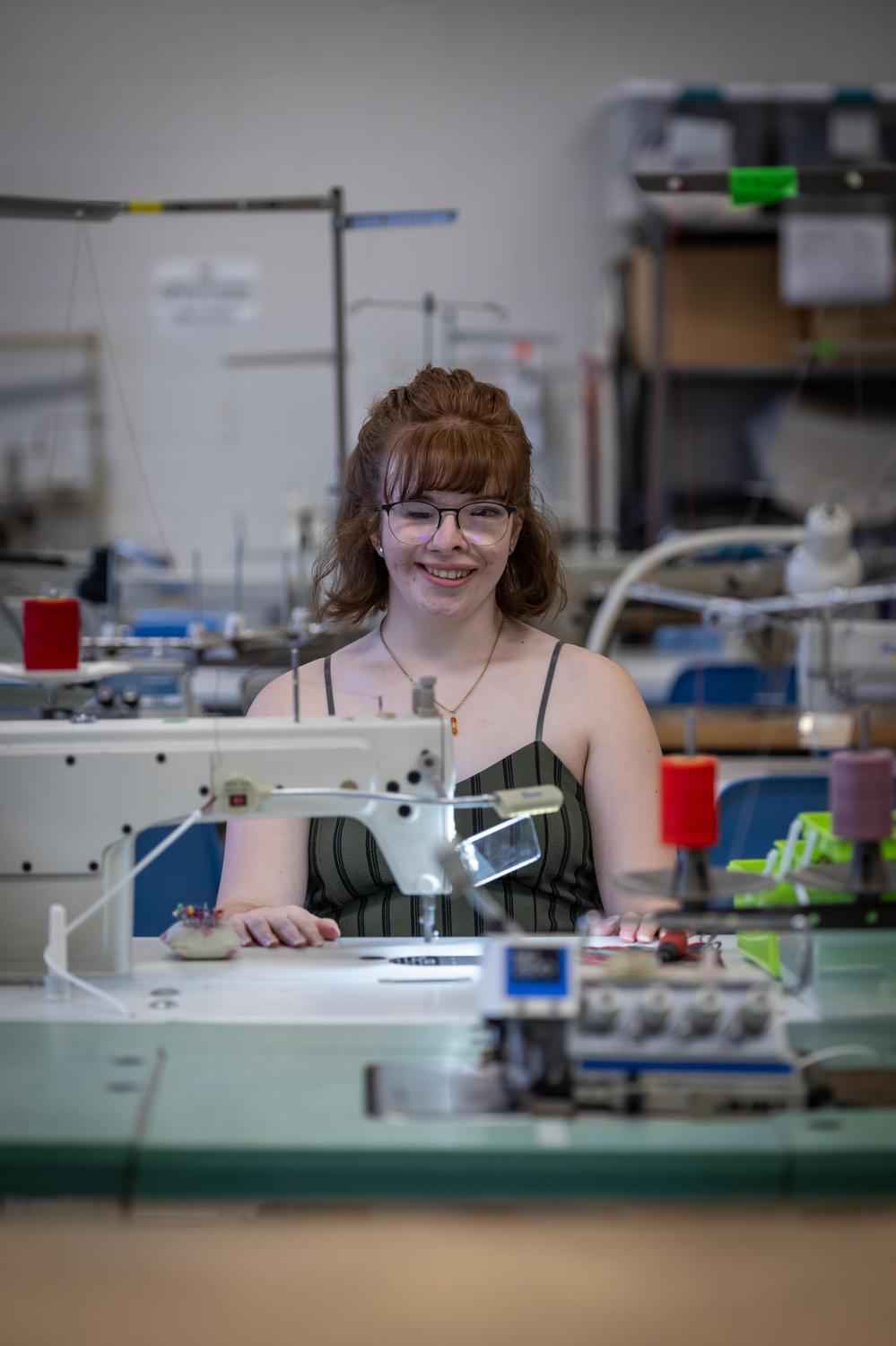 Young woman with glasses smiling in a sewing lab. She sits behind a sewing machine with red spools. The room is filled with equipment, creating a focused yet cheerful atmosphere.