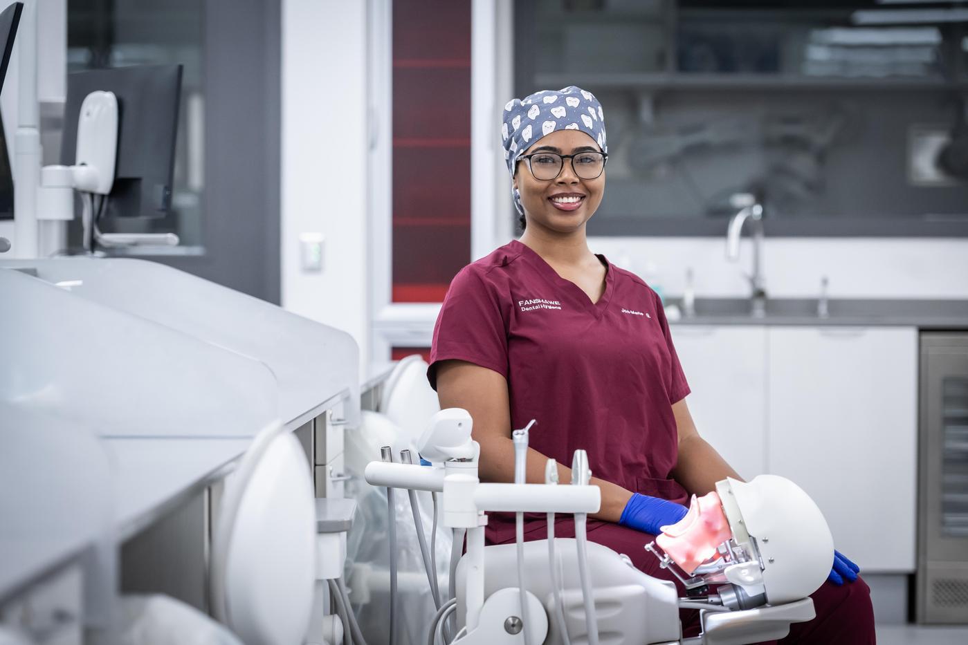In the School of Health Sciences at Fanshawe College's London Campus, someone wearing a burgundy scrub top and a surgical cap patterned with teeth sits in a dental chair, with a dental mannequin in their lap.