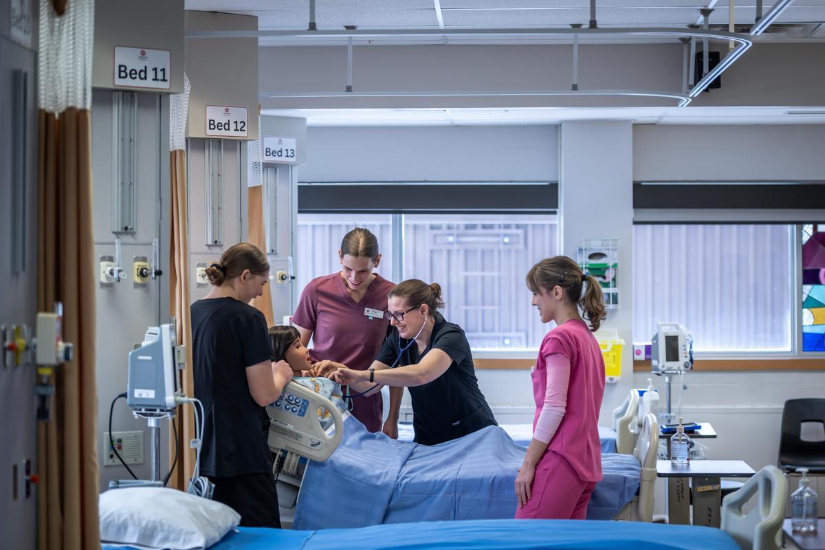 In a bright hospital room, four individuals in scrubs surround a patient in a bed with blue linens, one using a stethoscope