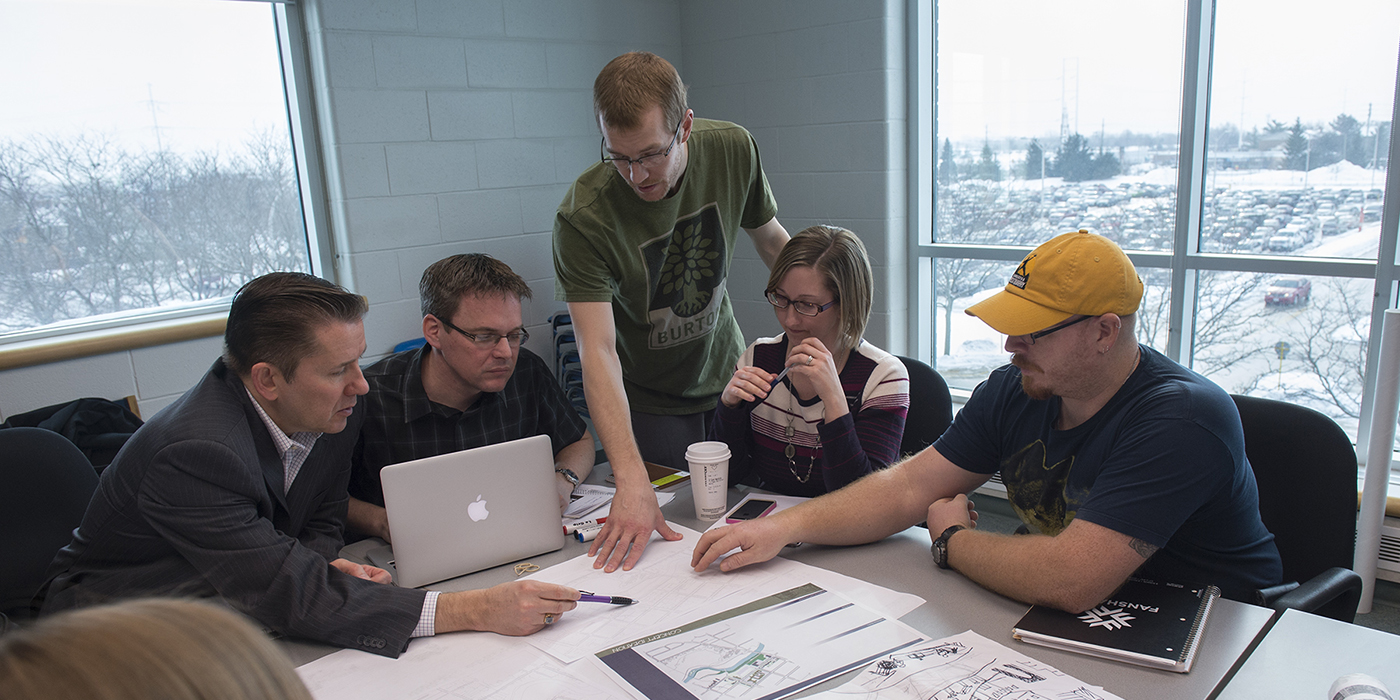 At the London Campus of Fanshawe College, a group gathers around a table covered with architectural drawings, a laptop, and notebooks, discussing plans with a large window overlooking a snowy parking lot in the background.