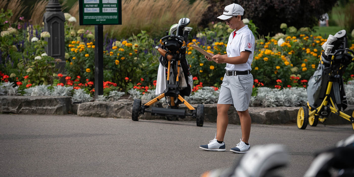 At a golf course, a person in a white polo shirt, grey shorts, and a white cap stands on a paved path, holding a clipboard, with golf bags and carts nearby, and a course status sign visible behind a flower bed.