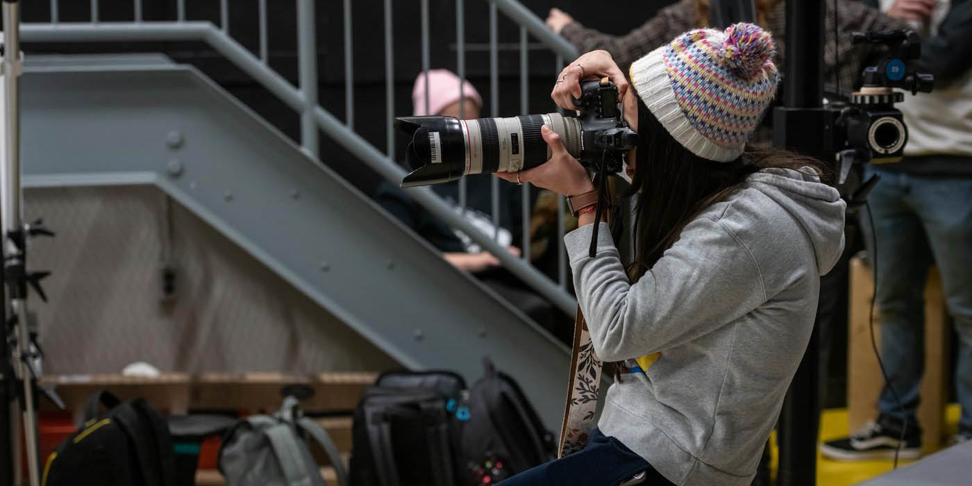 At the London Campus of Fanshawe College, someone wearing a colourful toque and grey hoodie sits on a wooden box, holding a camera with a long lens up to their eye, with a grey staircase and other people in the background.